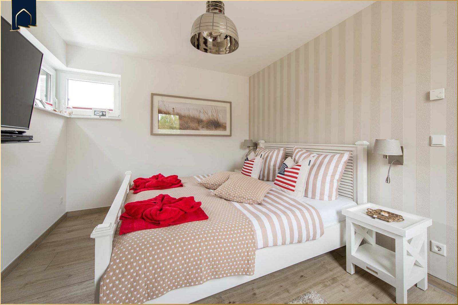 Schlafzimmer mit Bett, Nachttisch und Wandbild. An der Wand hängt ein Fernseher.