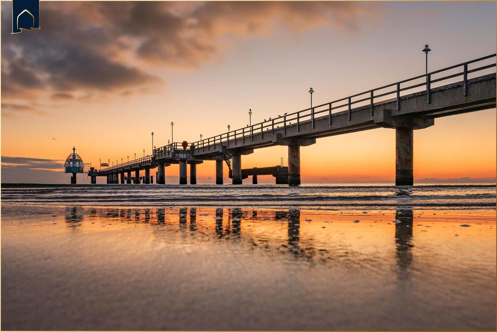 Eine Pier mit Laternen und einem Leuchtturm am Strand bei Sonnenuntergang.