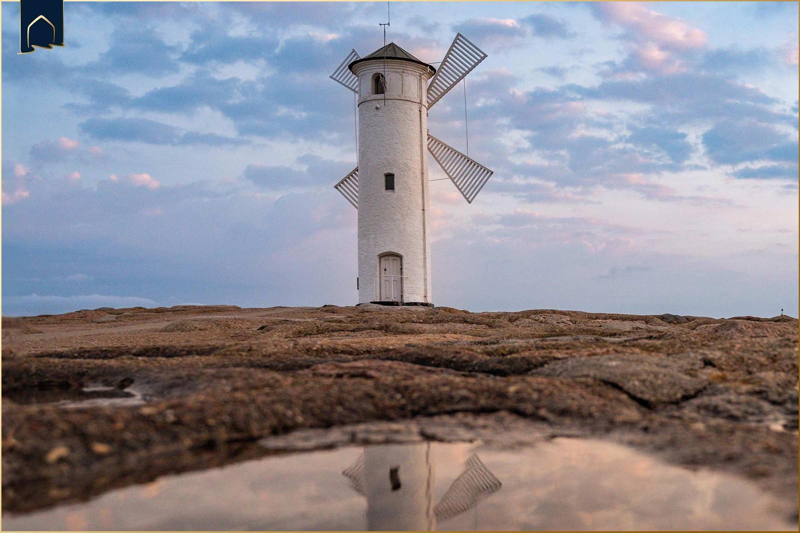 Weißer Windmühlenbau auf felsigem Terrain mit Reflexion im Wasser.