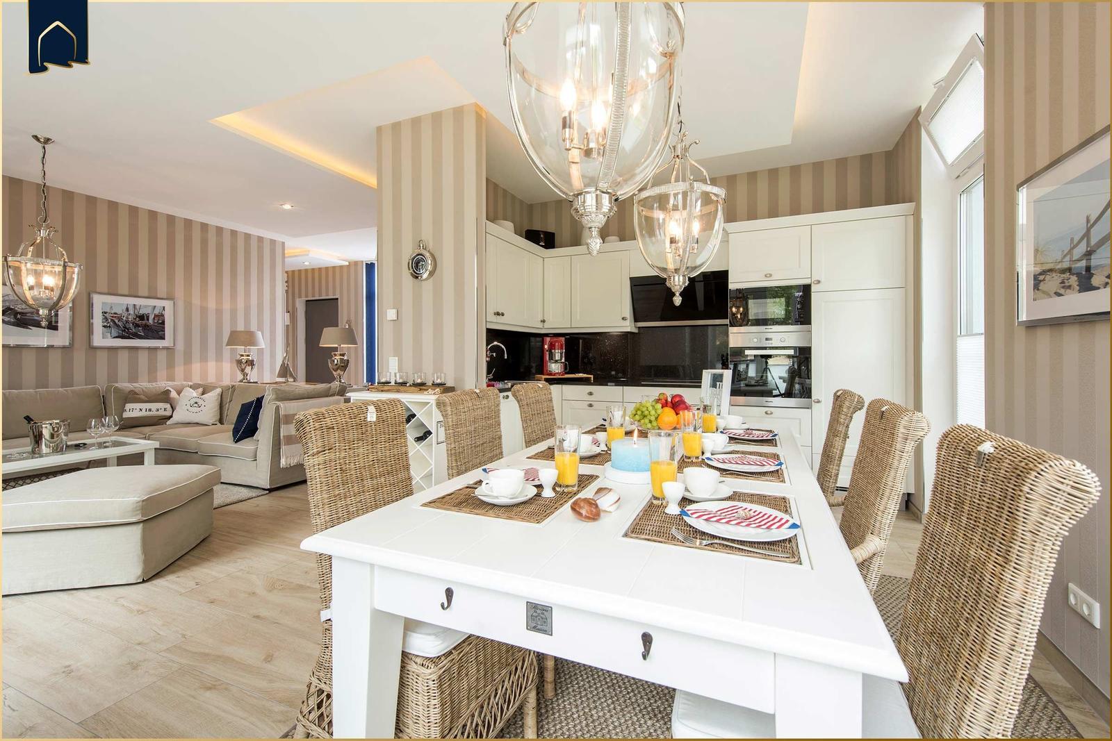 Esszimmer mit weißem Tisch und Rattanstühlen, Küche im Hintergrund, Wohnzimmer mit Sofa und Wandtapete.