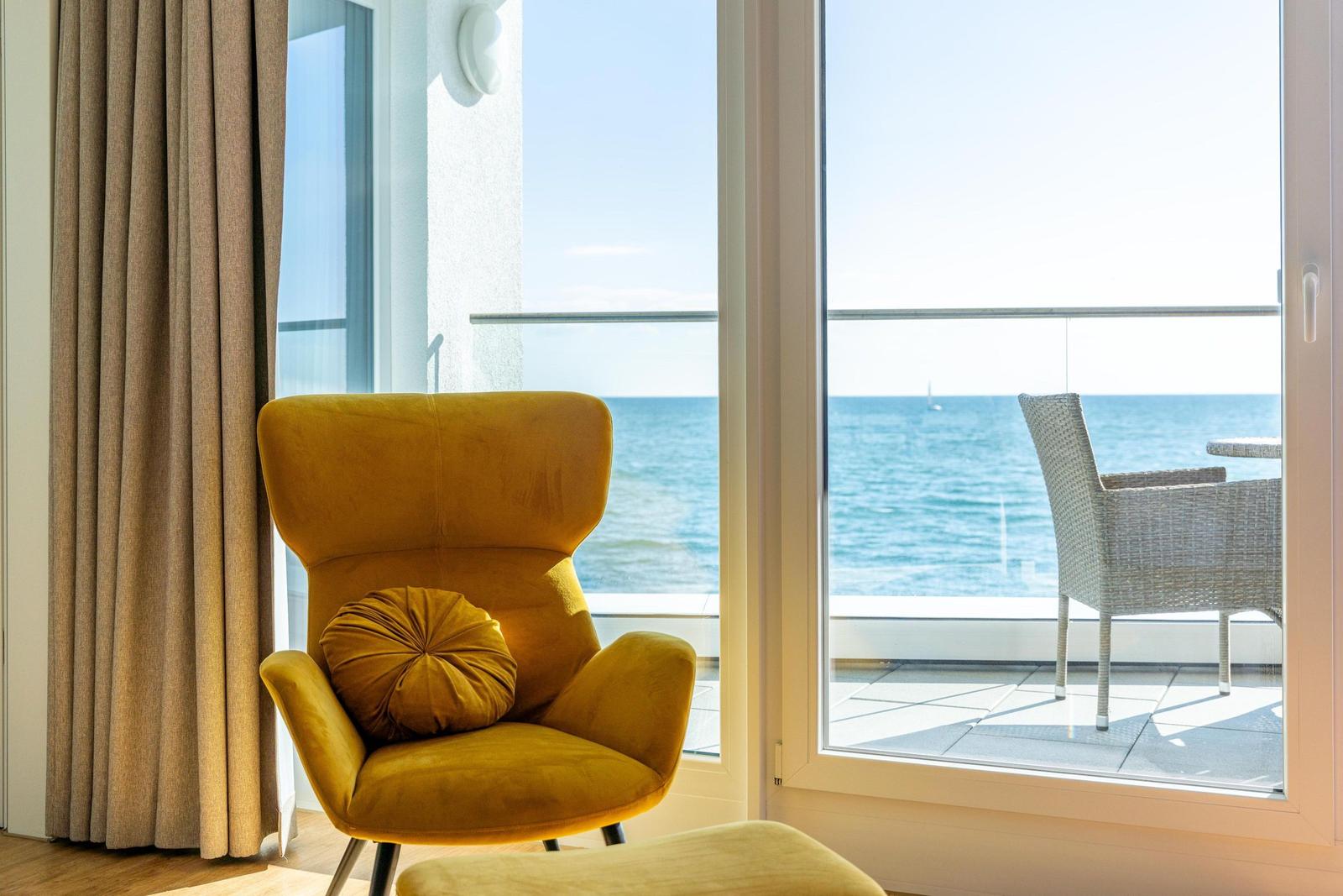 Gelber Sessel mit Kissen vor Fenster mit Meerblick und Balkonmöbeln.