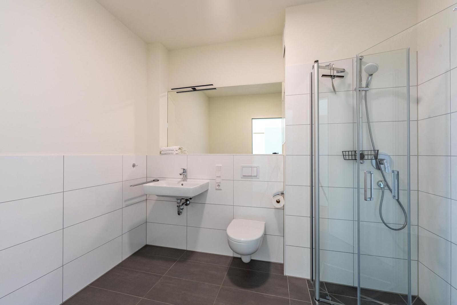 Badezimmer mit Dusche, Waschbecken und Toilette. Weiße Fliesen und graue Bodenfliesen.