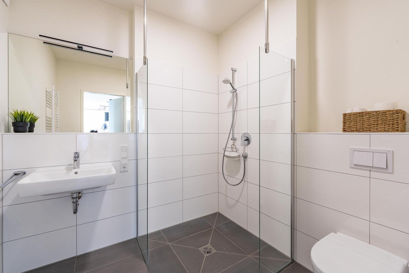 Badezimmer mit Dusche, Waschbecken und Toilette. Weiße Fliesen und graue Bodenfliesen.