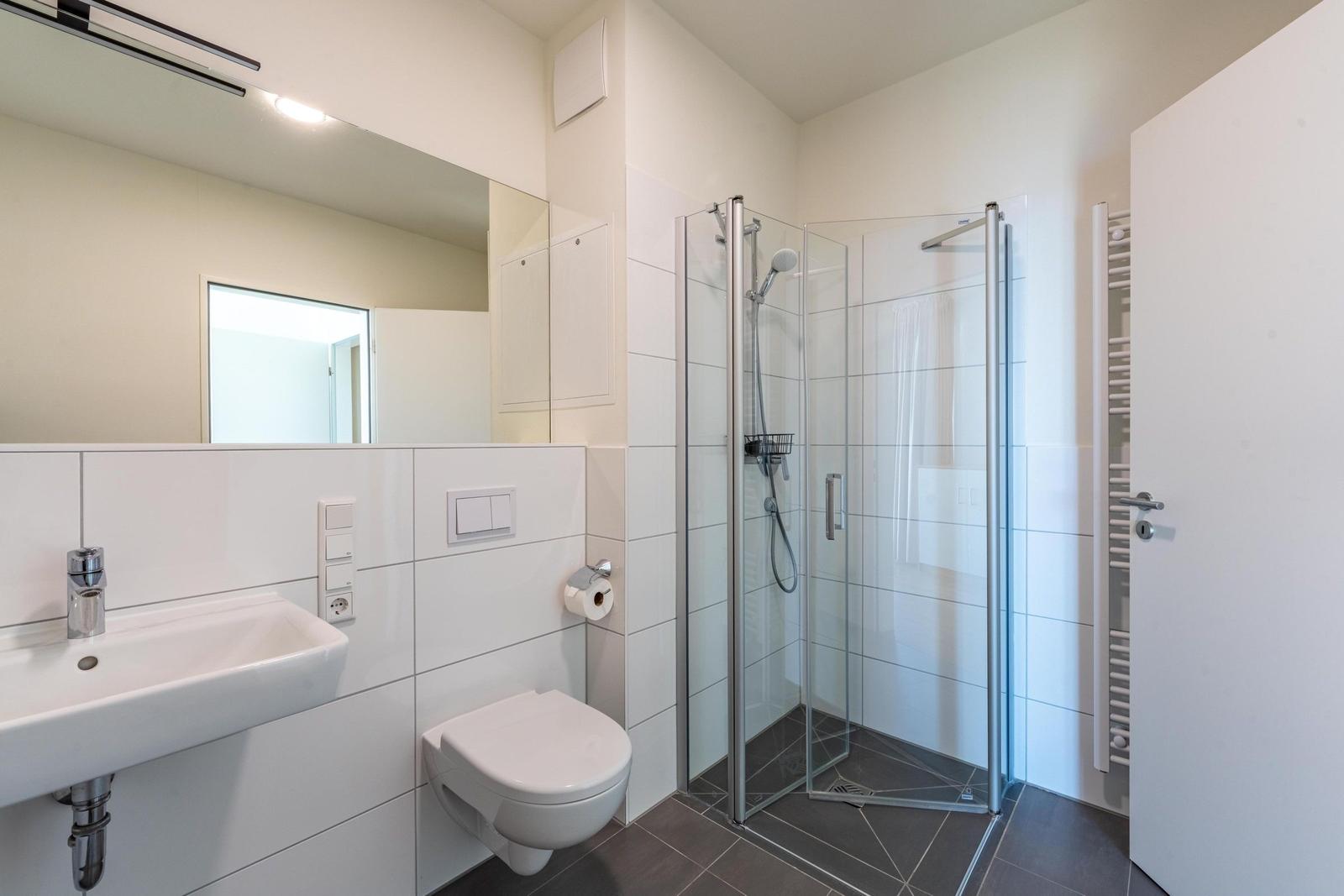 Badezimmer mit Dusche, Waschbecken und WC. Fliesen und Spiegel sind vorhanden.