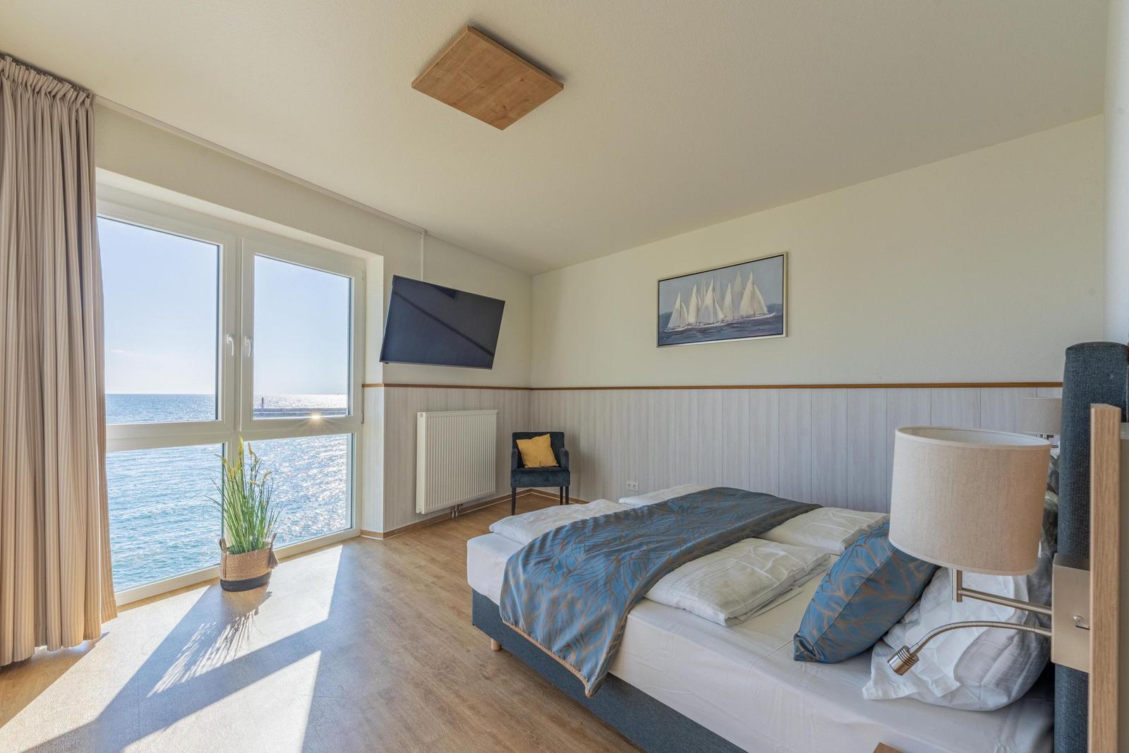 Schlafzimmer mit Blick aufs Meer, Bett, Fernseher und Wandbild mit Segelbooten.