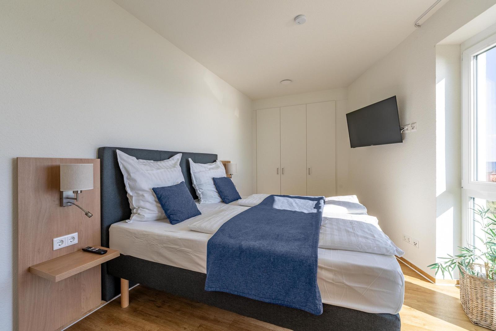 Schlafzimmer mit Doppelbett, Kleiderschrank und Wandfernseher. Fenster mit Pflanze.