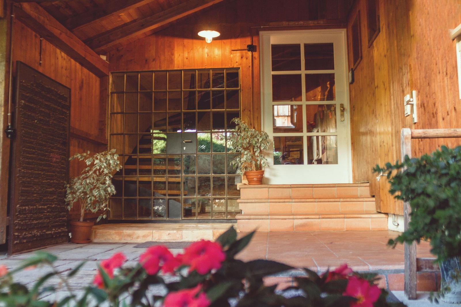 Eingangsbereich mit Holzwand, Glasfenster und Pflanzen. Tür führt ins Innere.