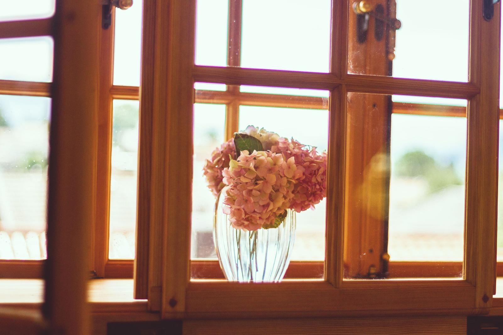 Ein Glas mit rosa Hortensien steht auf einem Fensterbrett in einem Holzfenster.