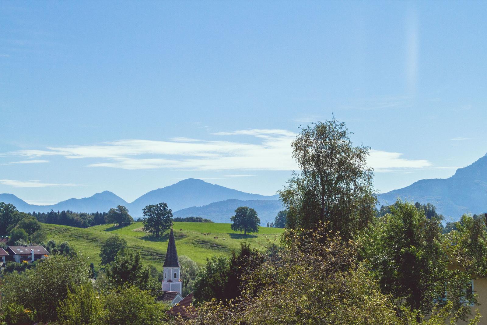 Weitläufiger Blick auf grüne Hügel mit Kirche und Bergen im Hintergrund unter blauem Himmel.