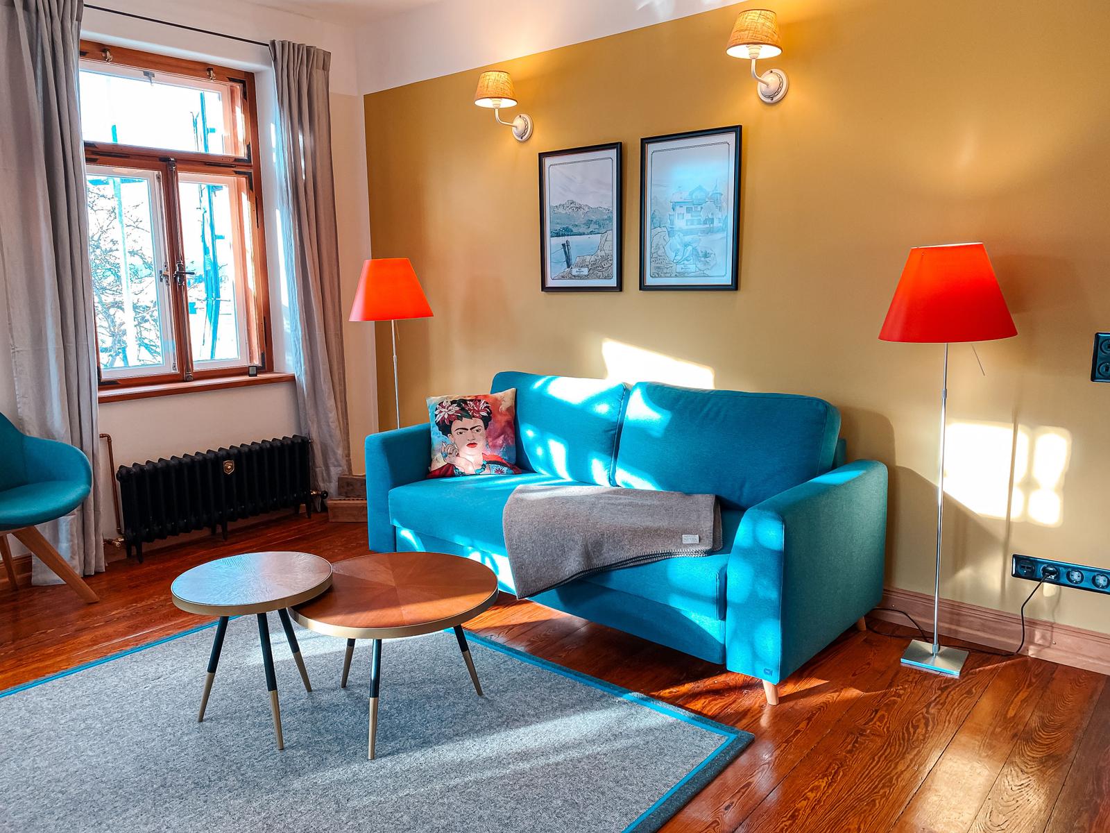 Ein Wohnzimmer mit blauem Sofa, zwei orangefarbenen Stehlampen und einem Fenster mit Blick nach draußen.