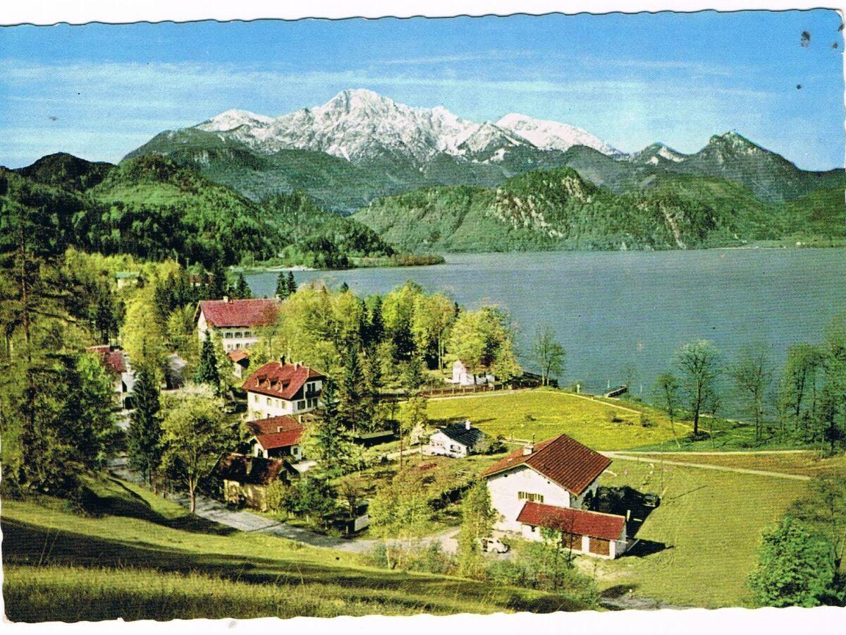 Haus mit rotem Dach am See, umgeben von Wäldern und Bergen mit Schnee.