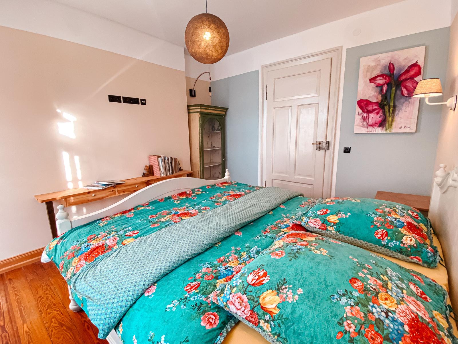 Schlafzimmer mit Bett, Wandbild und Holzboden. Decke mit floraler Bettwäsche.