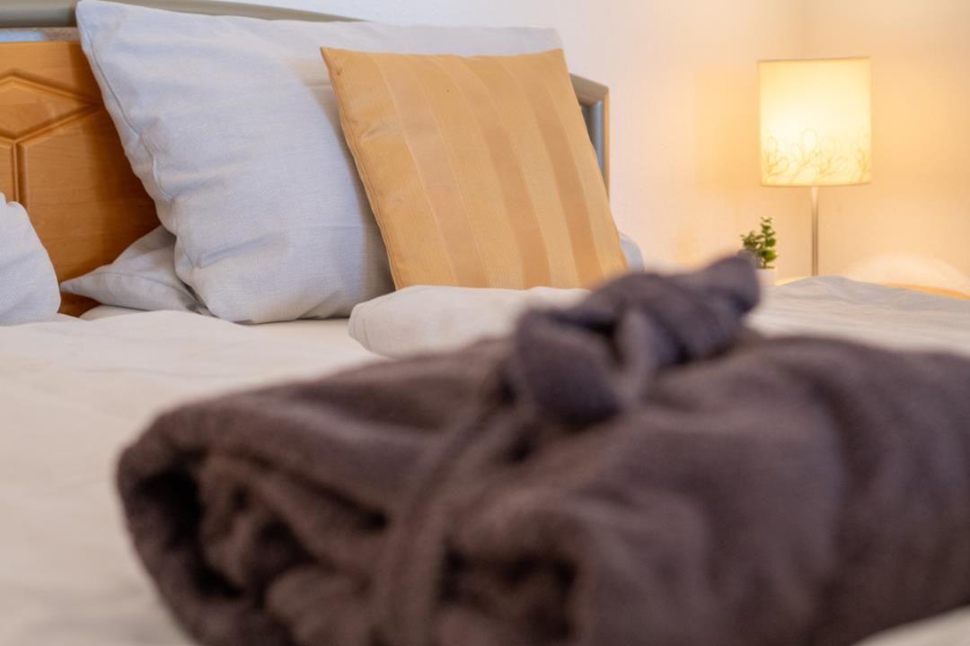 Ein Bett mit Kissen und einer Decke. Eine Lampe und eine kleine Pflanze sind sichtbar.