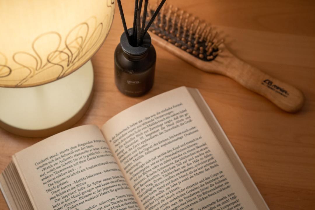 Ein offenes Buch liegt auf einem Holztisch neben einer Duftflasche und einem Haarbürsten.