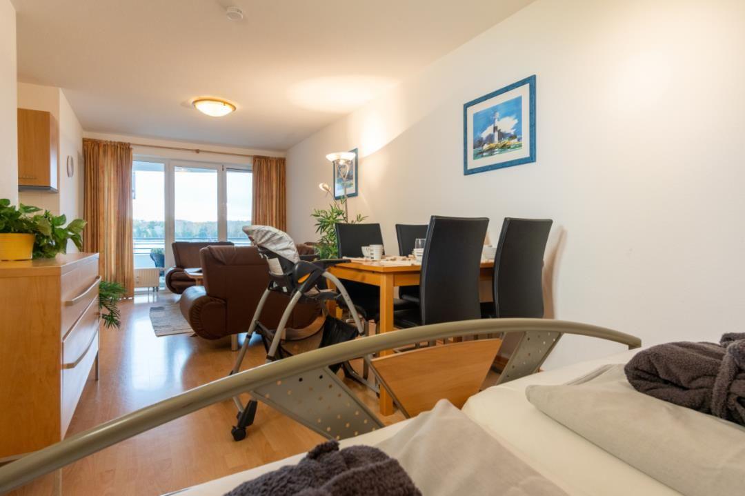 Wohn- und Essbereich mit Blick auf den See, Esstisch mit Stühlen und Bett im Vordergrund.