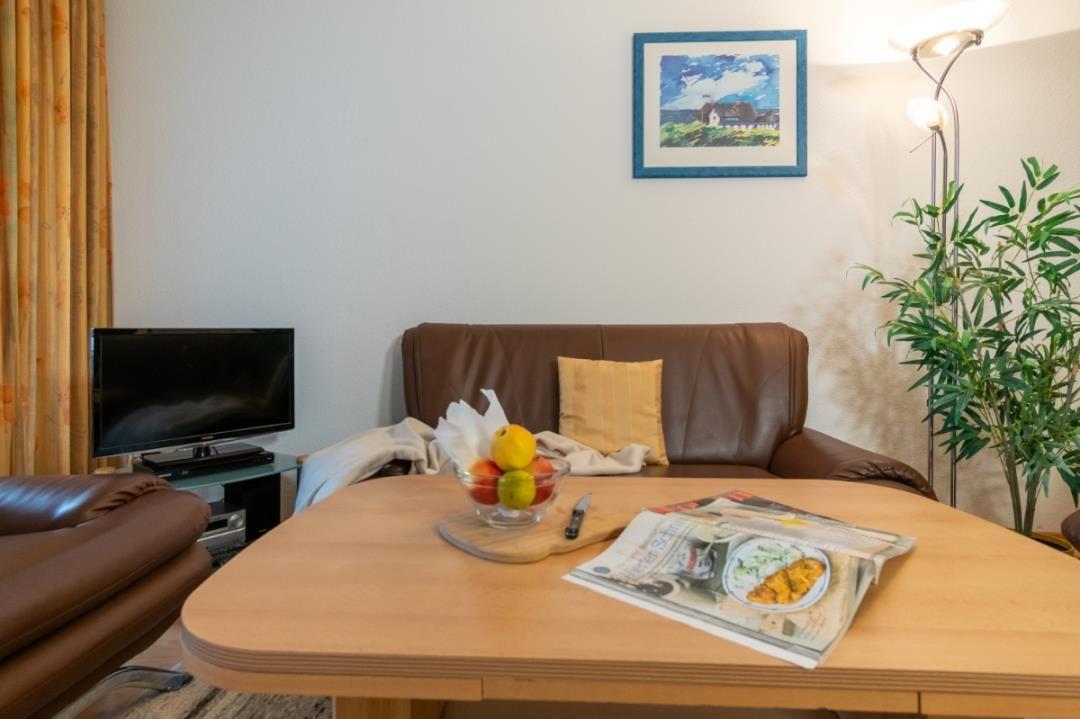 Ein Wohnzimmer mit braunem Sofa, Holztisch und Fernseher. Auf dem Tisch steht eine Obstschale.