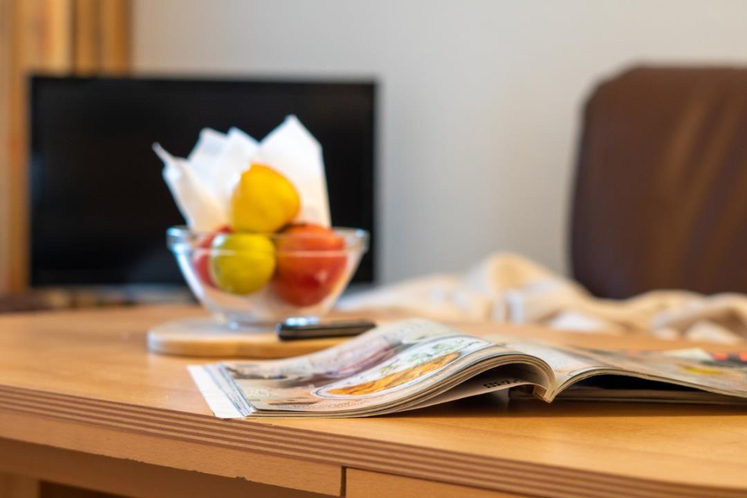 Holztisch mit offener Zeitschrift, Obstschale und Fernseher im Hintergrund.