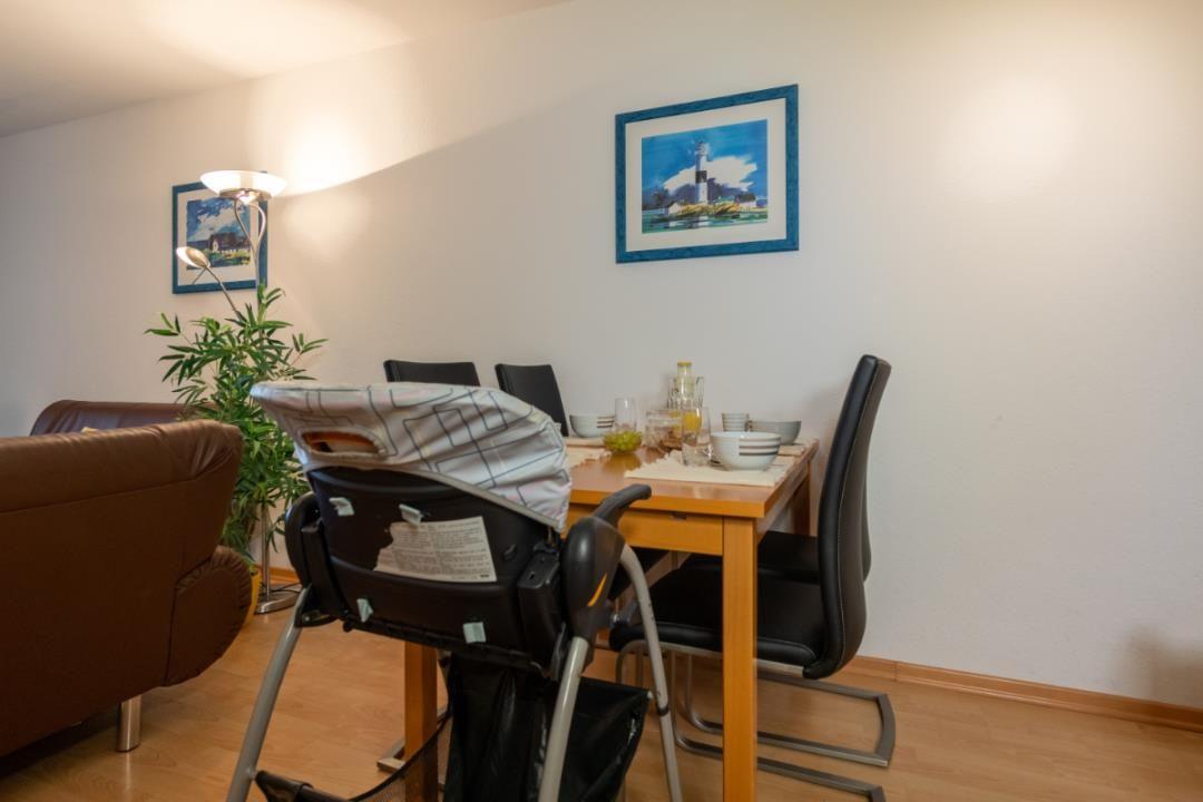 Essbereich mit Tisch, Stühlen und Hochstuhl. Wandbilder und Pflanze sind sichtbar.