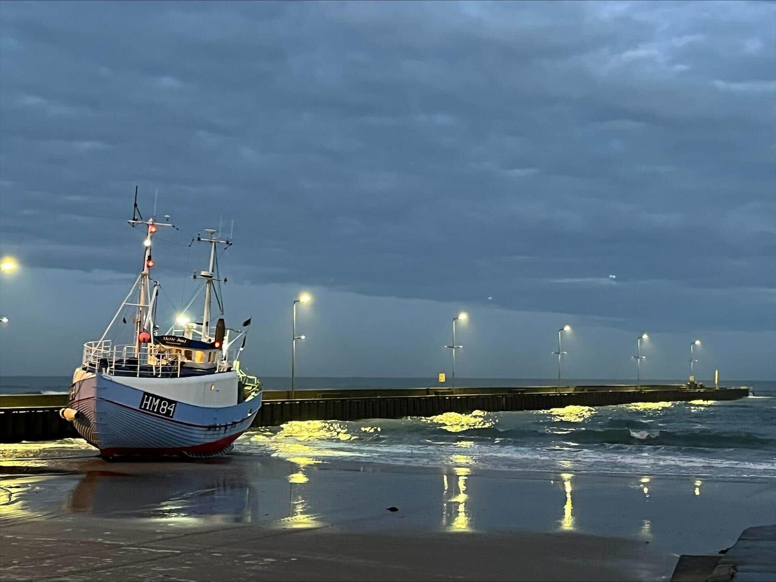Fishing boat HM84 rests on the beach at night. Waves break against the pier.