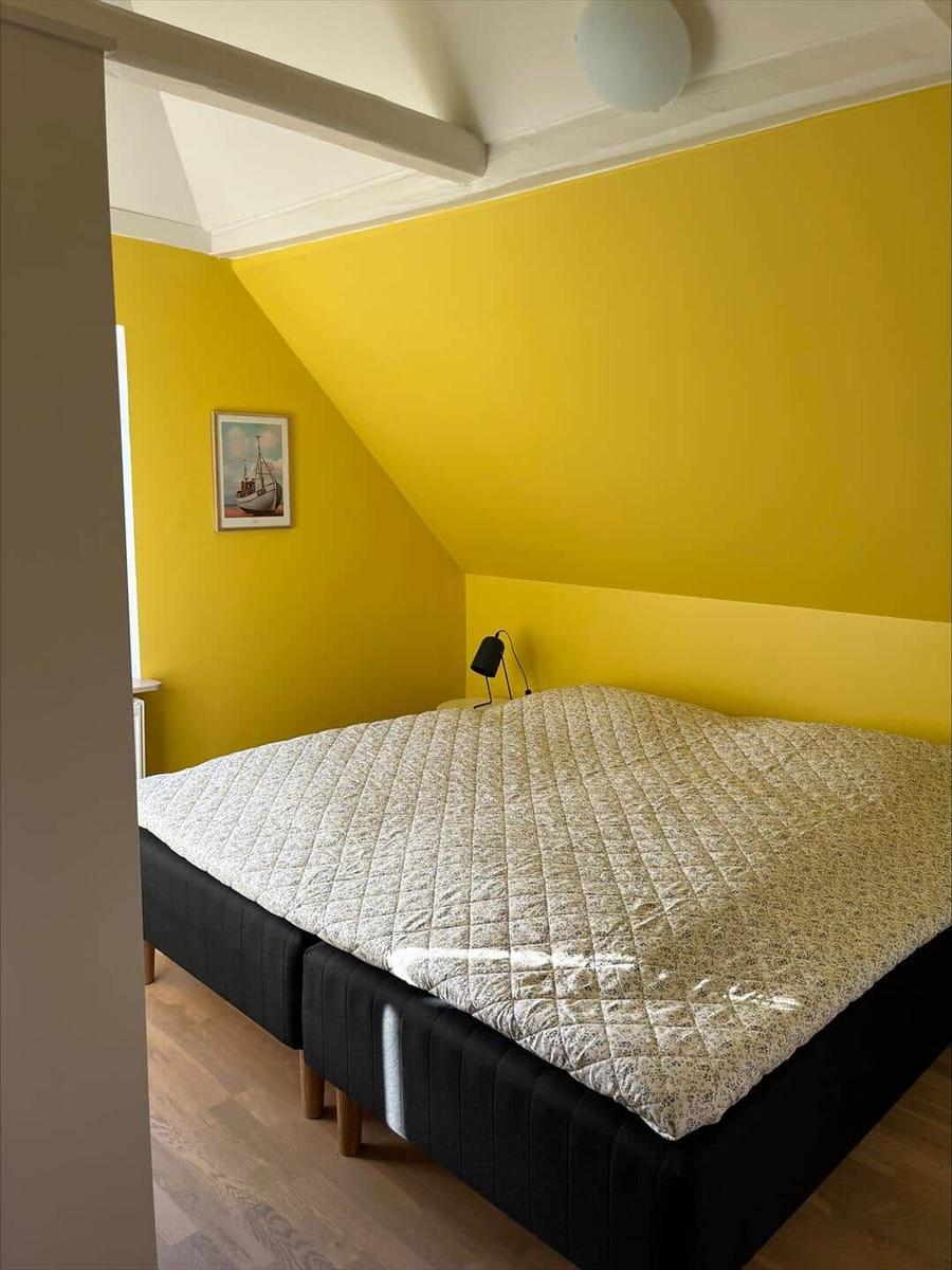 Bedroom with yellow walls, bed with patterned coverlet, and ceiling beams.