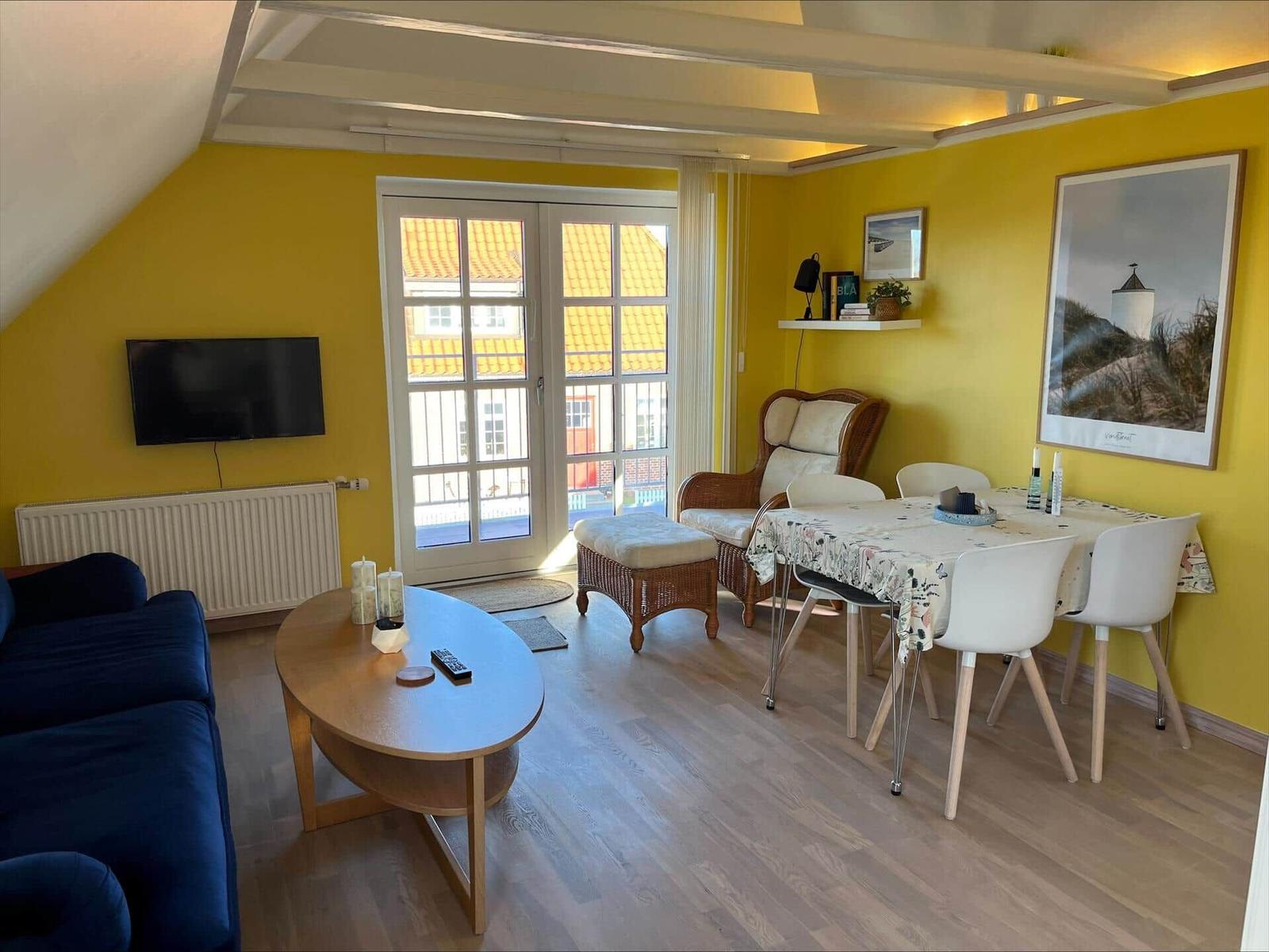 Living and dining area with yellow walls, TV, dining table, and balcony view.