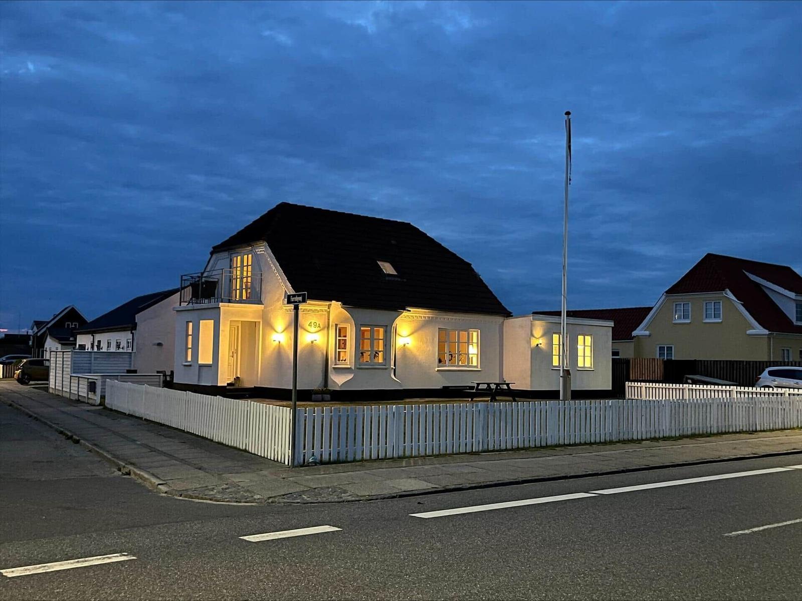 House with white facade, dark roof, and white fence. Windows are lit.