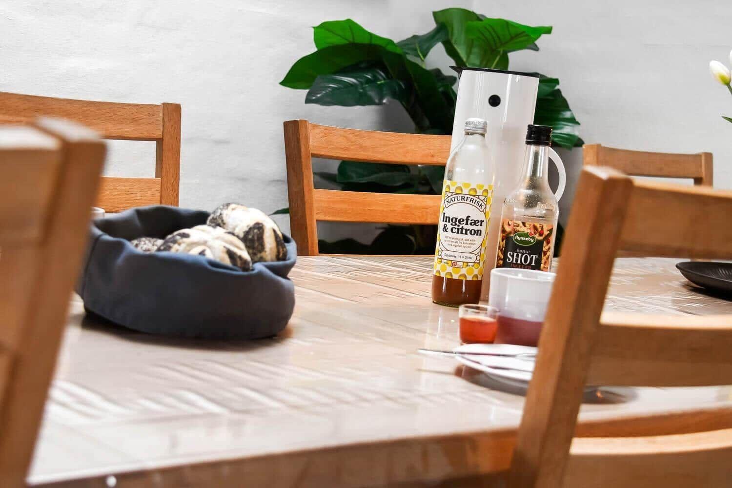 Wooden table with chairs, bread, drinks, and plant in background.