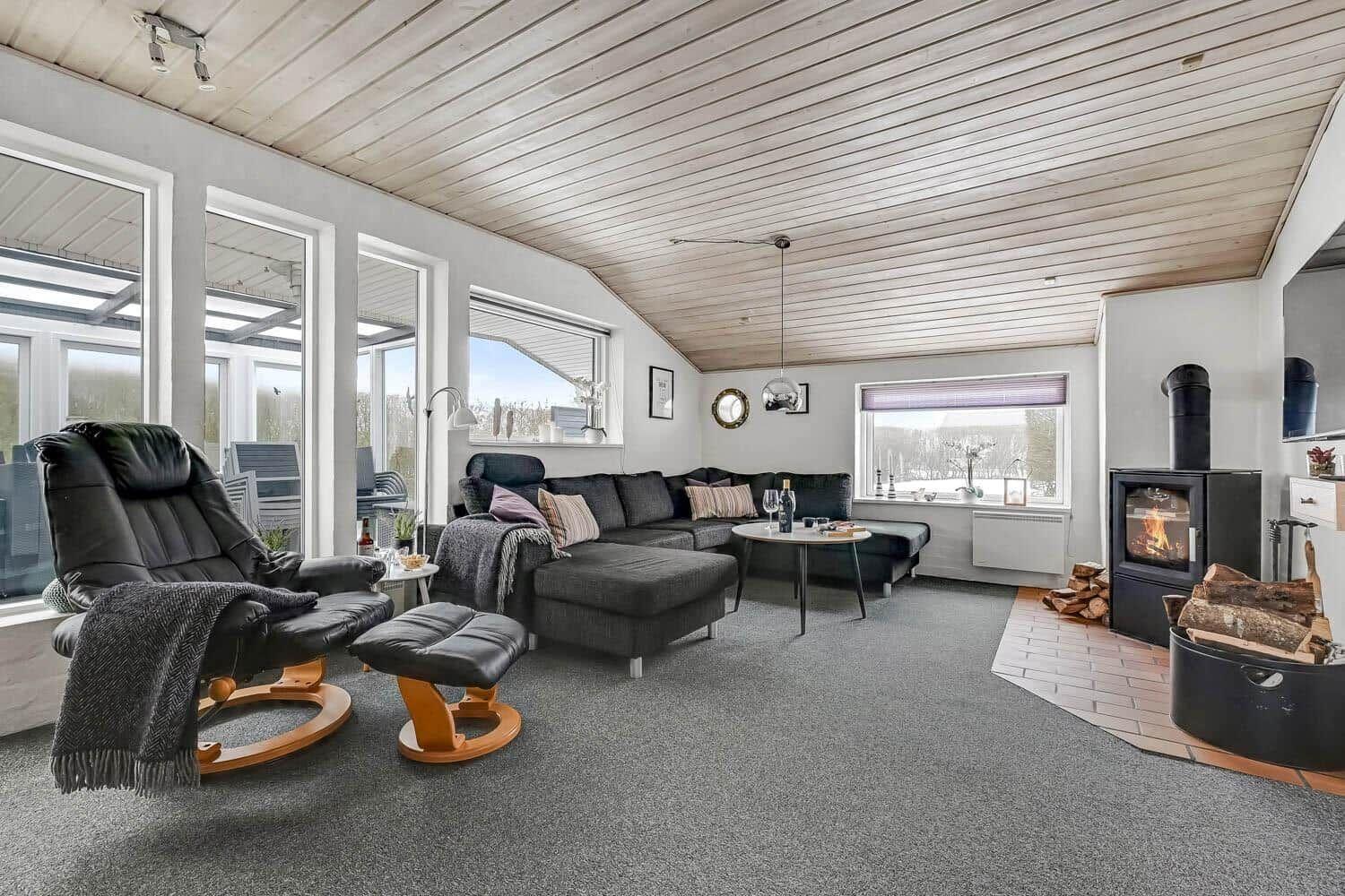 Living room with seating group, stove, and large window area. Wooden ceiling and carpeted floor.