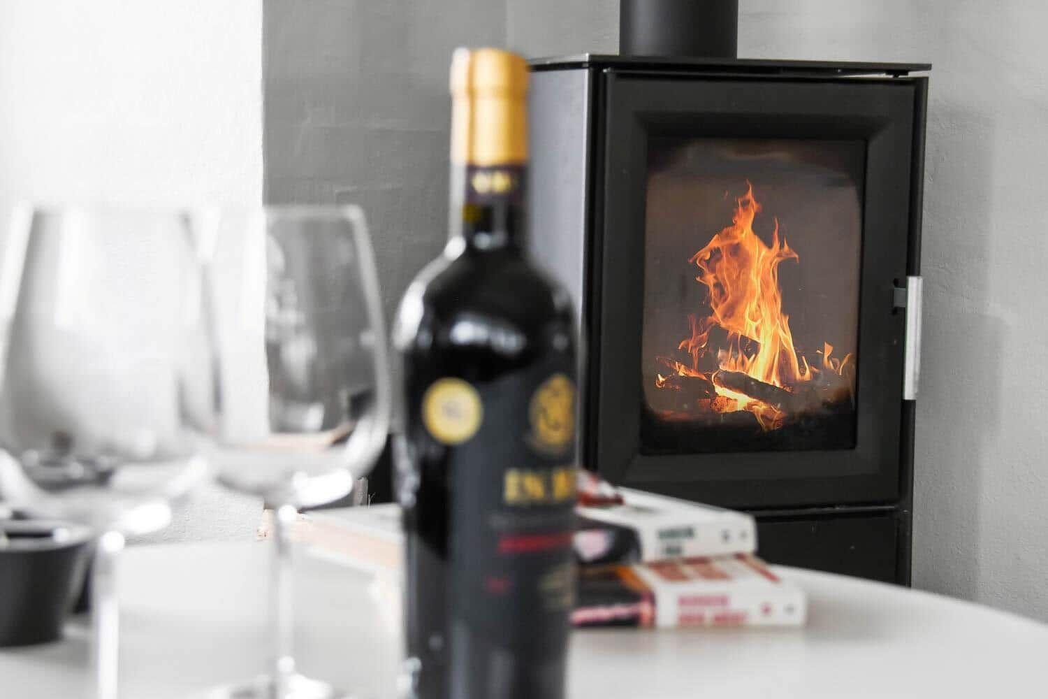 A table with a wine bottle and glasses in front of a burning wood stove.