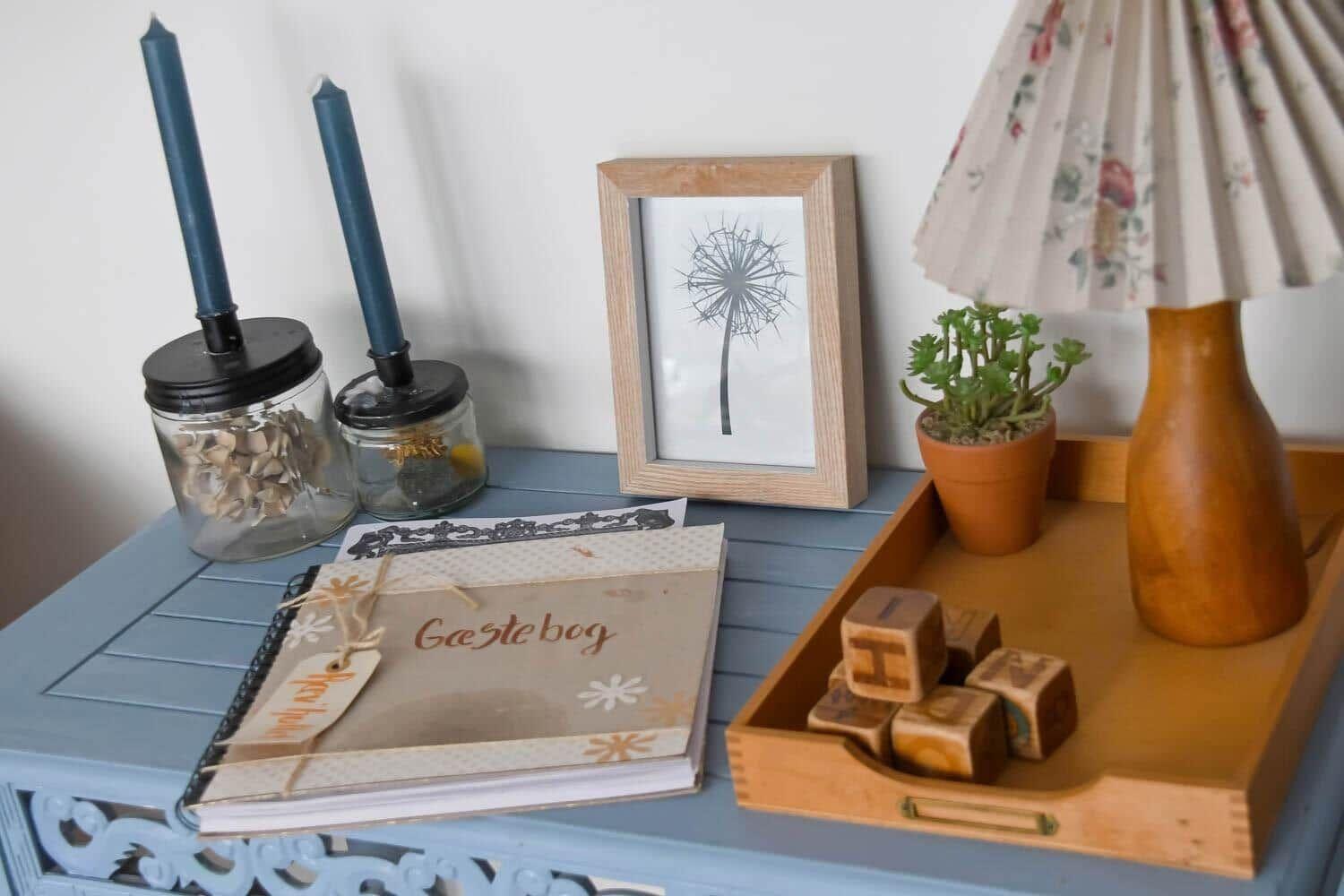 Auf einem blauen Tisch steht ein Gästebuch, eine Lampe, eine Pflanze und ein Bild mit einer Pusteblume.