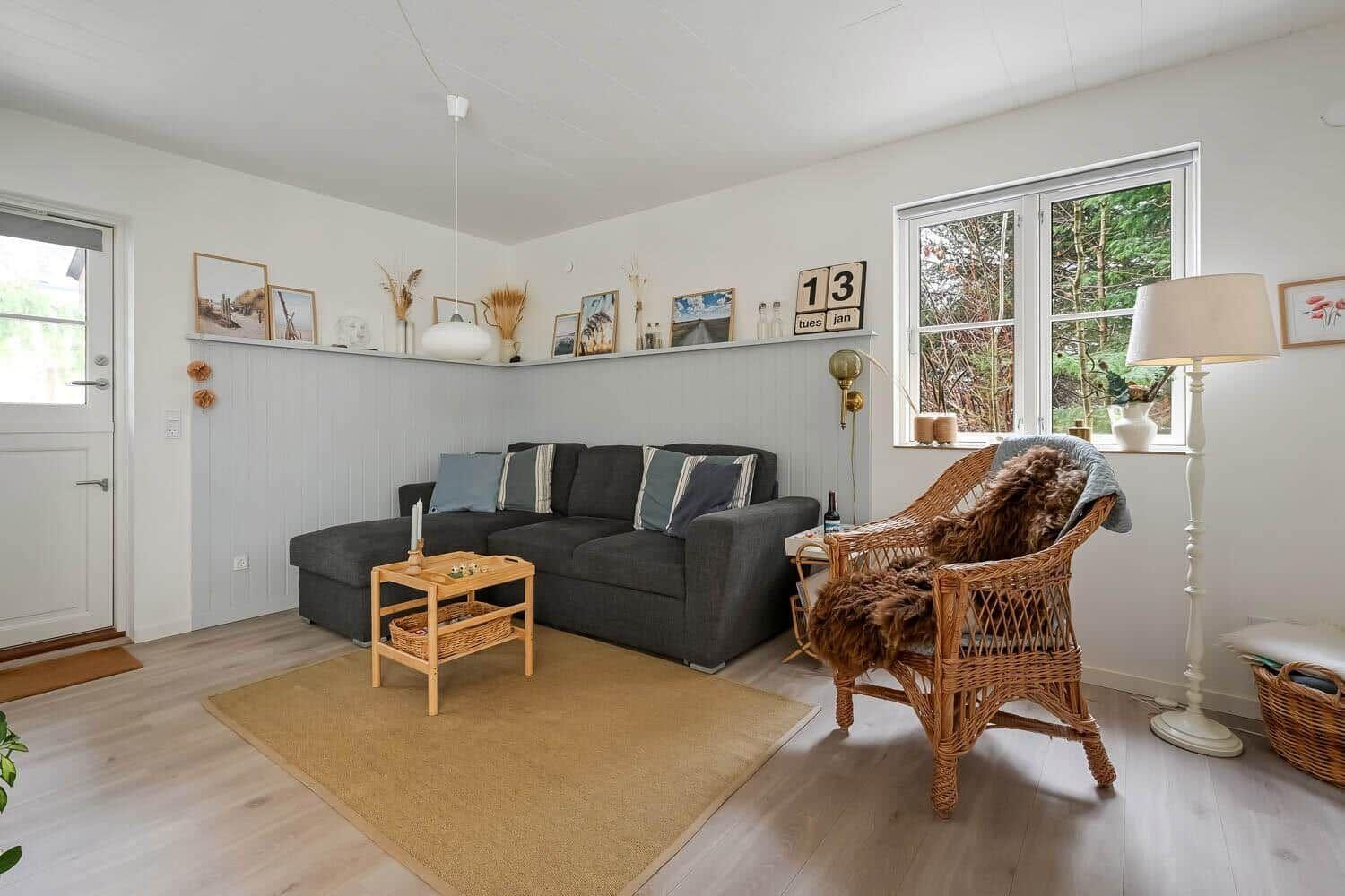 Wohnzimmer mit grauem Sofa, Bambusstuhl und Holzboden. Fenster mit Blick auf Wald.