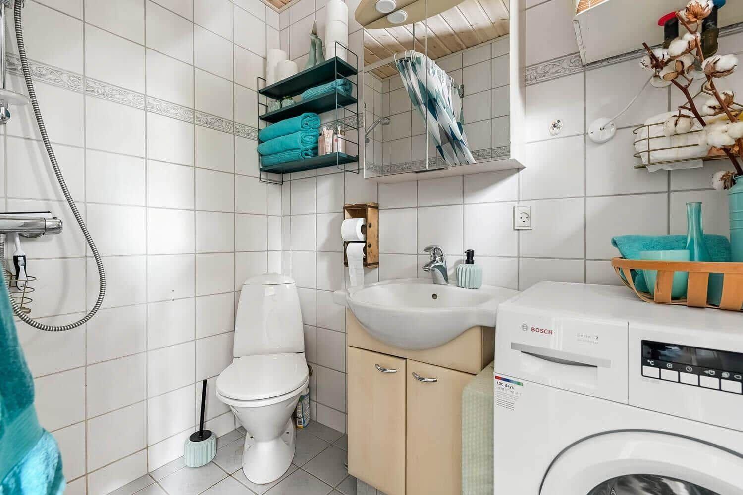 Badezimmer mit Toilette, Waschbecken und Waschmaschine. Weiße Fliesen und blau-grüne Handtücher.