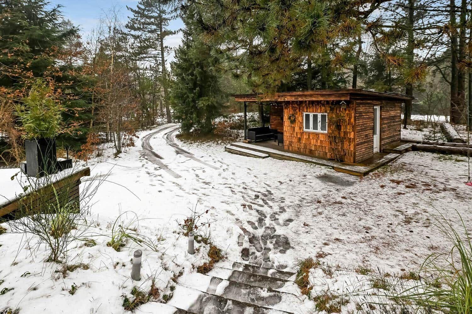 Holzhütte mit Terrasse im Schnee, umgeben von Bäumen und einem schneebedeckten Weg.