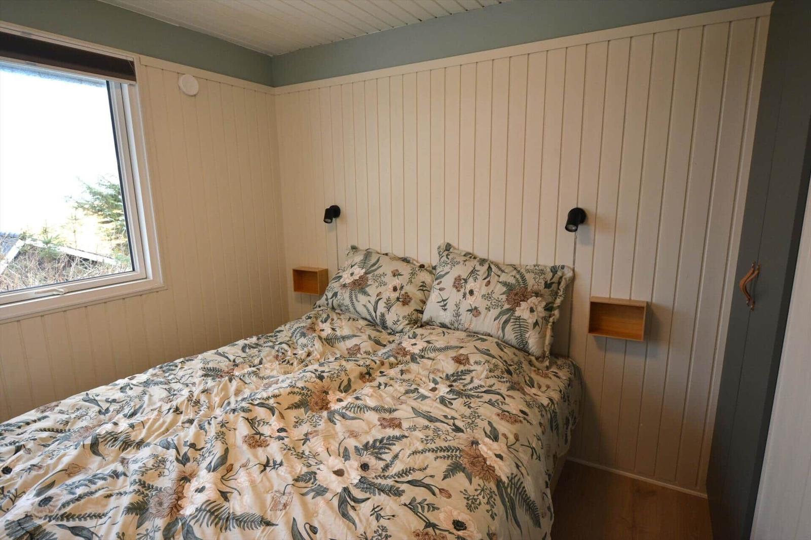 Schlafzimmer mit Bett, Fenster und Holzwandverkleidung. Zwei Nachttische und Wandlampen.