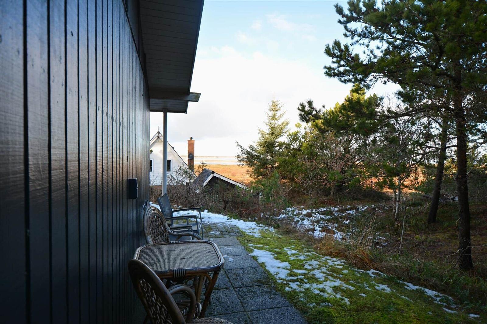 Außenbereich mit Tisch und Stühlen neben einem dunklen Holzhaus. Gras und Schnee im Hintergrund.