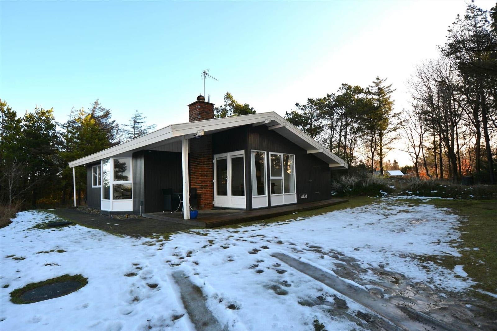 Ein Haus mit dunkler Fassade, weißem Dach und Fenstern. Schnee bedeckt den Boden. Bäume umgeben das Grundstück.
