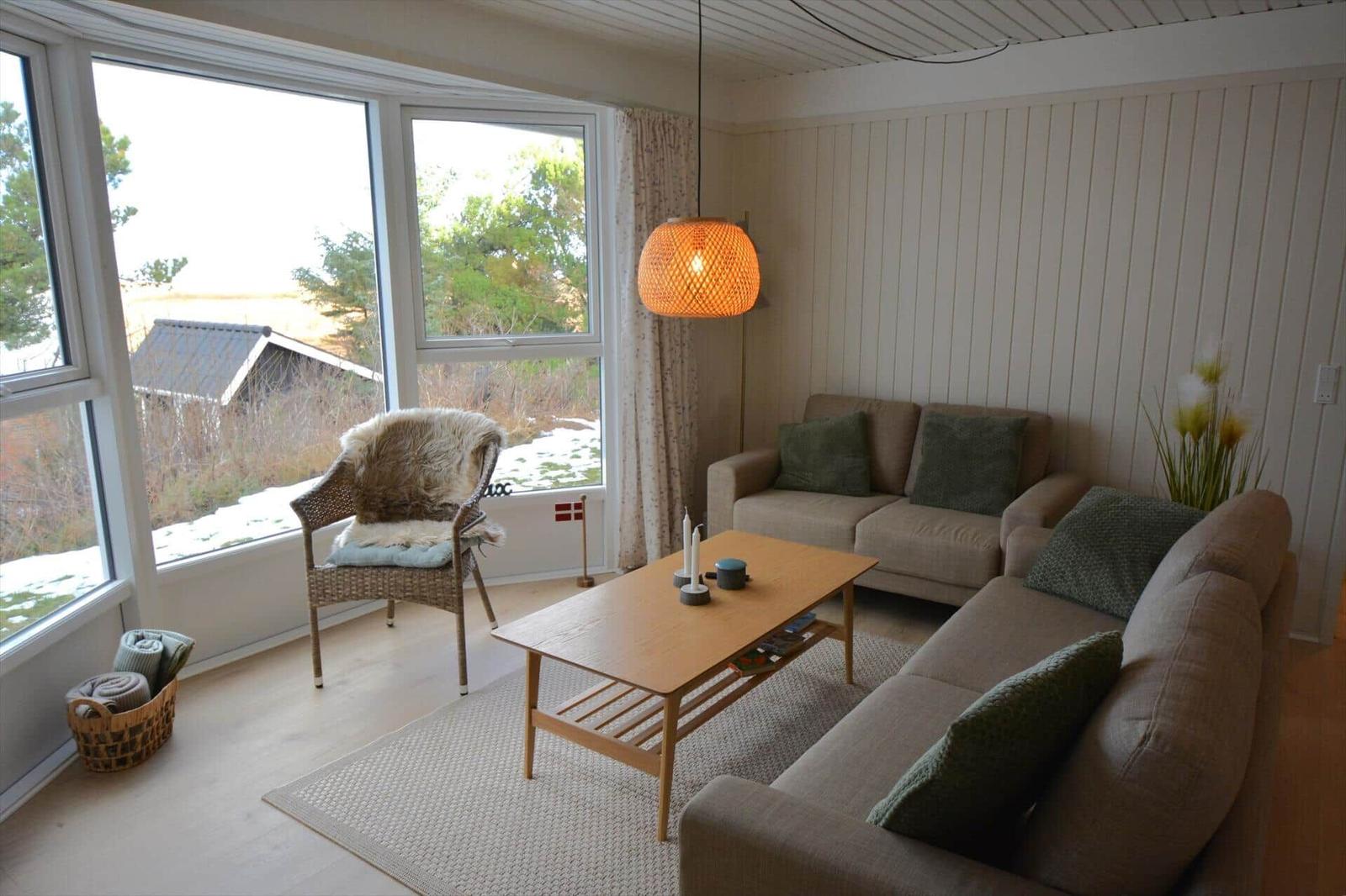 Wohnzimmer mit Sofa, Sessel und Tisch. Große Fenster mit Blick auf Außengelände.