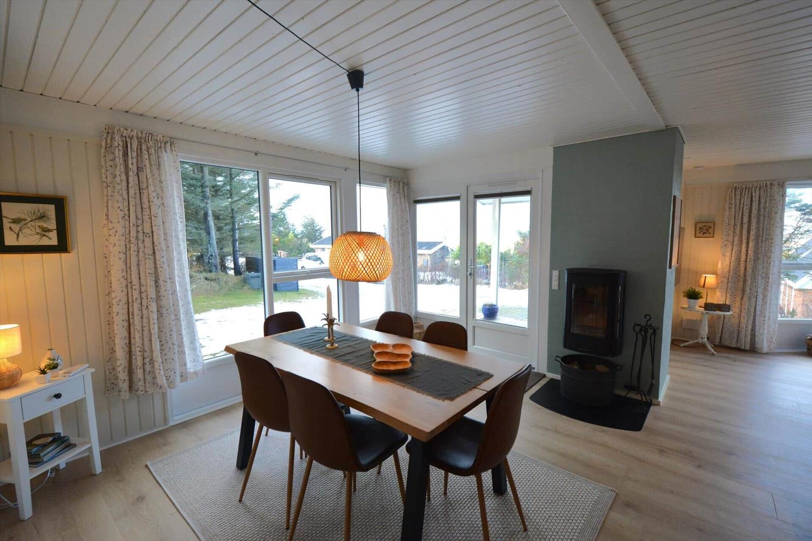 Esszimmer mit Holztisch, Stühlen und Kamin. Große Fenster und helle Wände.
