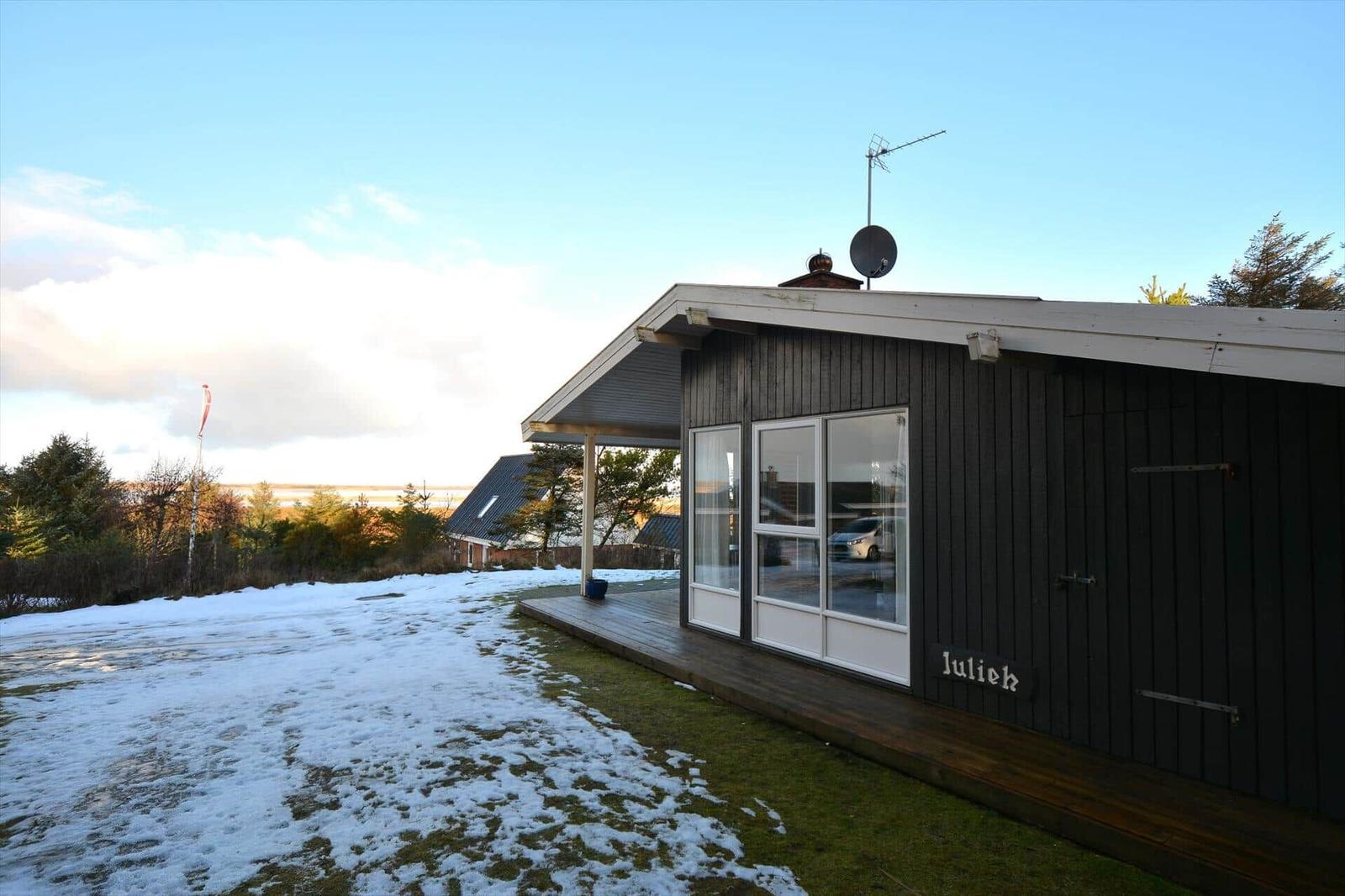 Schwarzes Holzhaus mit Terrasse und Schneebedecktem Grundstück. Satellitenantenne und Fenster sichtbar.