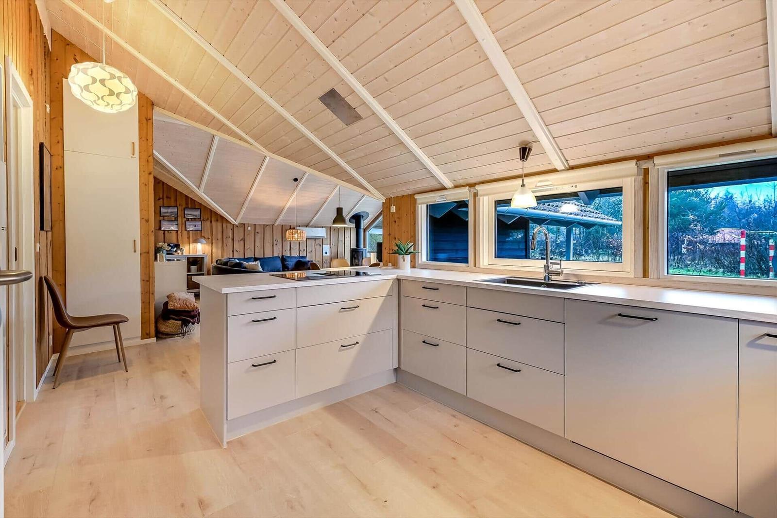 Küche mit Holzdecke, Fenstern und Arbeitsplatten. Wohnbereich im Hintergrund sichtbar.