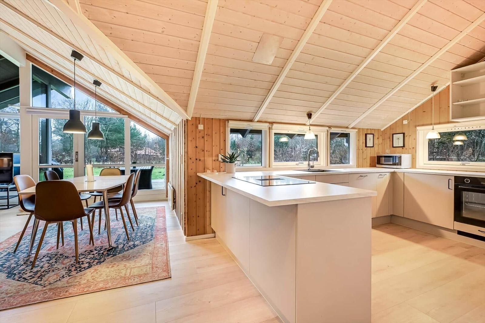 Küche mit Essbereich, Holzdecke und großen Fenstern. Weiße Arbeitsplatten und moderne Geräte.