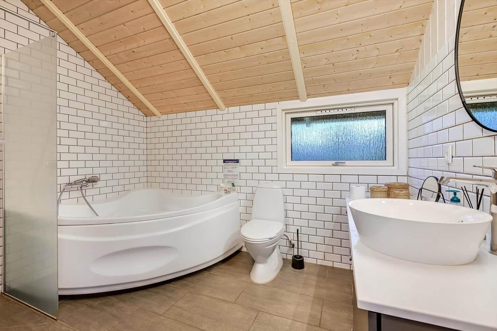 Badezimmer mit Badewanne, Toilette und Waschbecken unter Holzdecke mit weißen Fliesen.