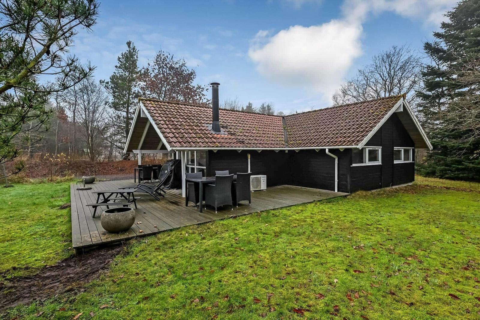 Schwarze Holzhütte mit Terrasse, Grill und Garten. Hintergrund: Wald und Himmel.