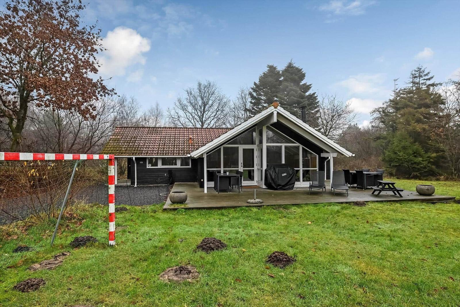 Haus mit Holzterrasse, Garten und Grillplatz. Umgeben von Bäumen und Grasfläche.