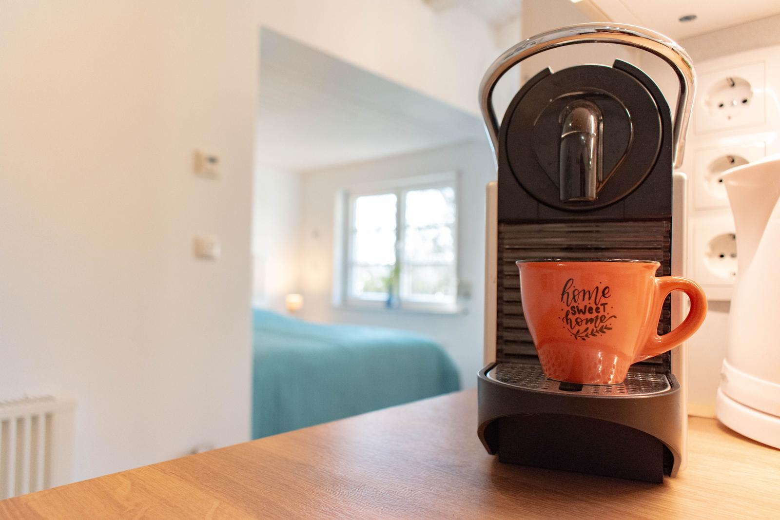 Kaffeemaschine mit Tasse auf Küchenarbeitsplatte. Im Hintergrund ist ein Schlafzimmer mit Bett und Fenster sichtbar.