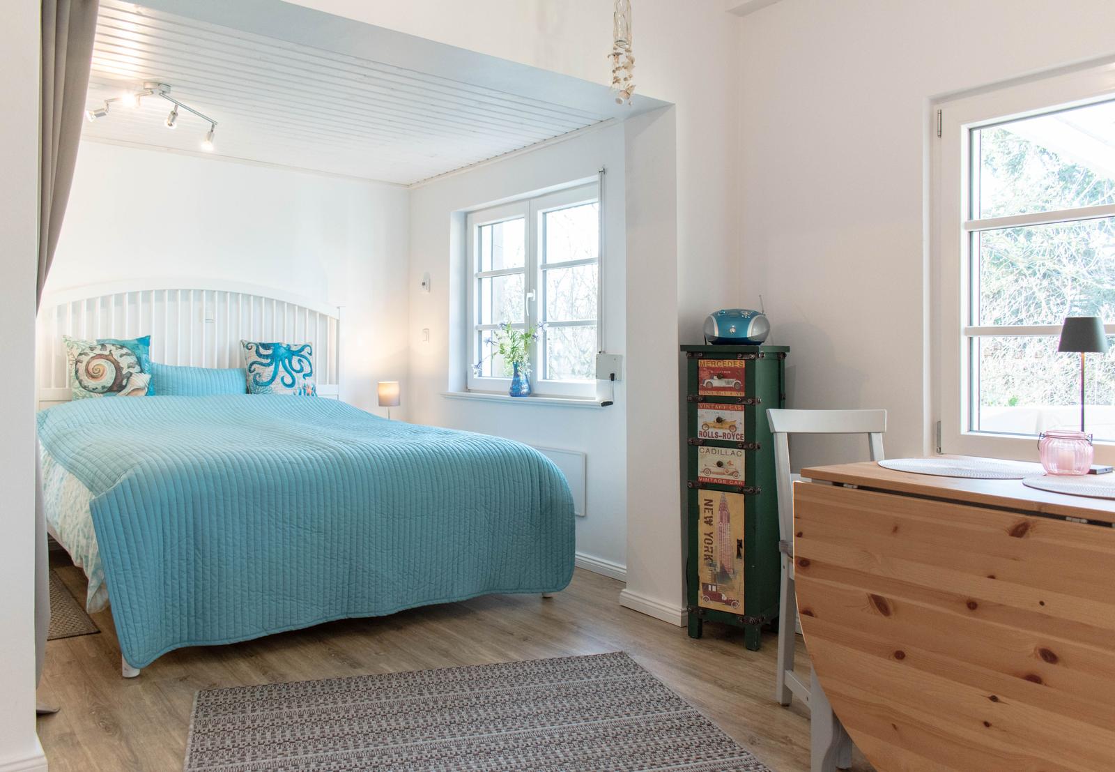 Schlafzimmer mit Bett, Schreibtisch und Fenstern. Holzboden und weiße Wände.