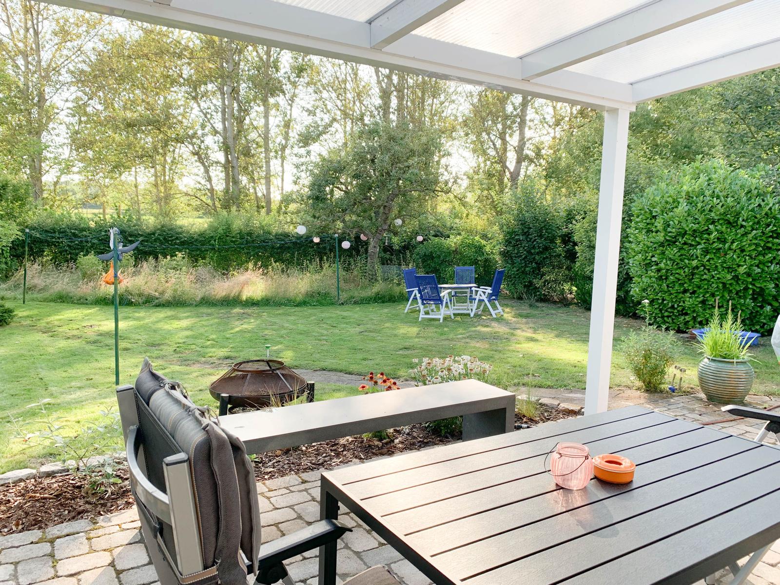 Terrasse mit Tisch, Stühlen und Grill unter Pavillon. Grünfläche mit Gartenmöbeln im Hintergrund.