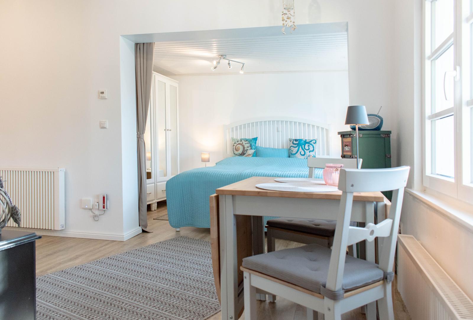 Ein Raum mit Tisch und Stühlen. Durchgehendes Fenster und Blick in Schlafzimmer mit Bett.