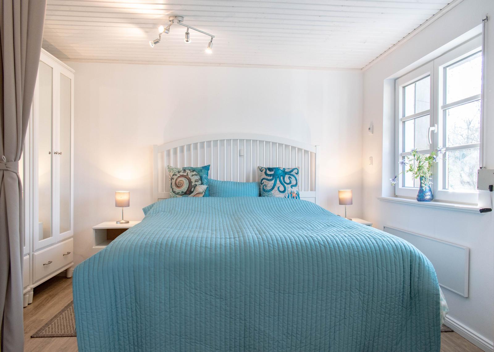 Schlafzimmer mit Bett, Nachttischen und Fenster. Decke weiß, Bett mit blauer Bettdecke.