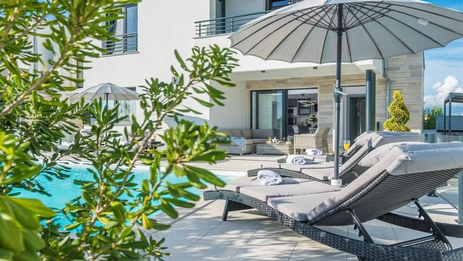 Pool area with lounge chairs and umbrellas. Modern house in the background.